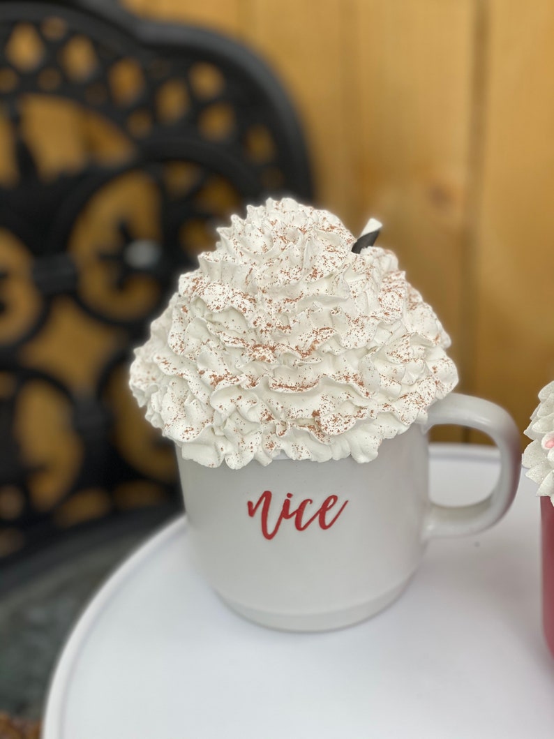 May include: White ceramic mug with a whipped cream topping and a sprinkle of cocoa powder. The mug has the word "nice" written on it in red.