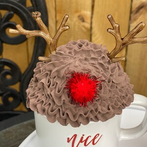 May include: White ceramic mug with brown whipped cream and reindeer antlers. The mug has the word "nice" written on it in red lettering. A red pom-pom sits on top of the whipped cream.