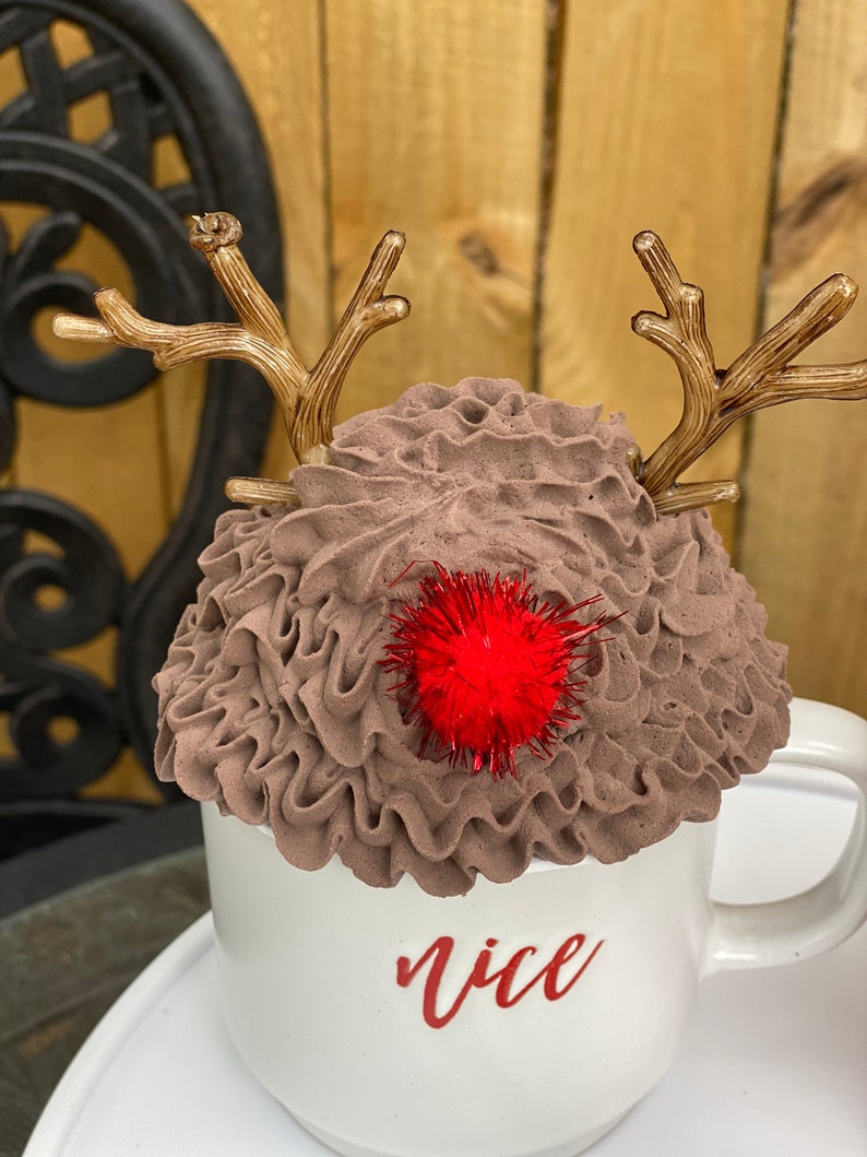 May include: White ceramic mug with brown whipped cream and reindeer antlers. The mug has the word "nice" written on it in red lettering. A red pom-pom sits on top of the whipped cream.