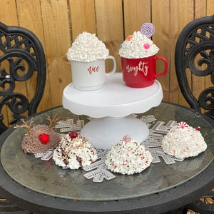 May include: Two white mugs with whipped cream and candy toppings. One mug says "nice" and the other says "naughty". Five additional whipped cream desserts with various toppings are on a black table with a snowflake design.