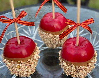 May include: Three red caramel apples with a brown caramel coating and a sprinkle of granola on top. Each apple is on a stick and has a red and orange striped bow.