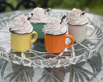 May include: Four miniature ceramic mugs with whipped cream and chocolate sprinkles on top. The mugs are in different colors: yellow, green, orange, and white. The mugs are sitting on a clear glass tray.