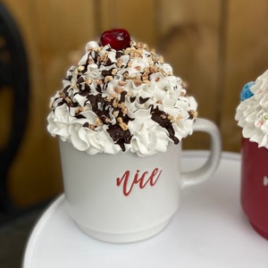 May include: White ceramic mug with a whipped cream and chocolate topping, a red cherry on top, and the word "nice" written in red on the side.