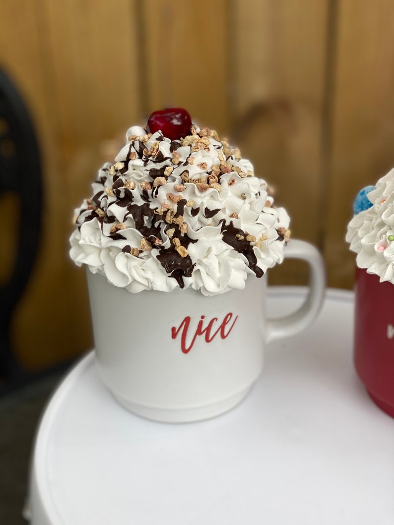 May include: White ceramic mug with a whipped cream and chocolate topping, a red cherry on top, and the word "nice" written in red on the side.