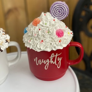 May include: Red ceramic mug with white whipped cream, colorful candies, and a purple swirl lollipop on top. The mug has the word "naughty" written on the front in white.