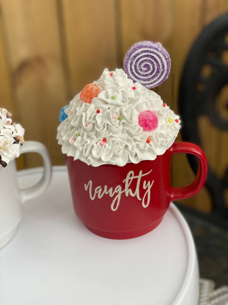 May include: Red ceramic mug with white whipped cream, colorful candies, and a purple swirl lollipop on top. The mug has the word "naughty" written on the front in white.