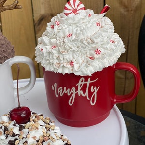 May include: Red ceramic mug with white whipped cream and a candy cane on top. The mug has the word "naughty" written on it in white.