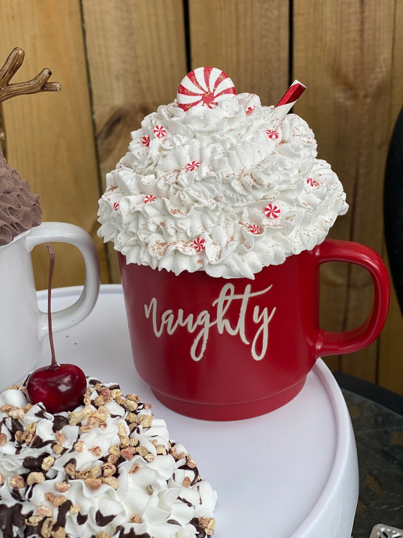 May include: Red ceramic mug with white whipped cream and a candy cane on top. The mug has the word "naughty" written on it in white.