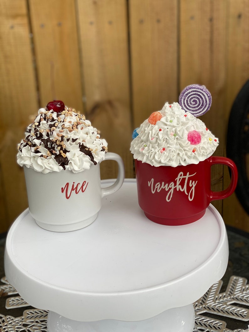 May include: Two ceramic mugs decorated to look like sundaes. The mug on the left is white with whipped cream, chocolate sauce, nuts, and a cherry on top. It says "nice" on the side. The mug on the right is red with whipped cream, sprinkles, candy, and a swirl lollipop on top. It says "naughty" on the side.