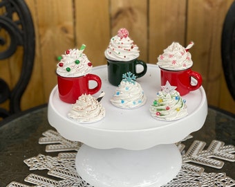 May include: A white cake stand with six miniature Christmas-themed hot chocolate mugs and cupcakes. The mugs are red and green with white frosting and sprinkles. The cupcakes are white with red and green sprinkles. The stand is on a silver snowflake-shaped doily.
