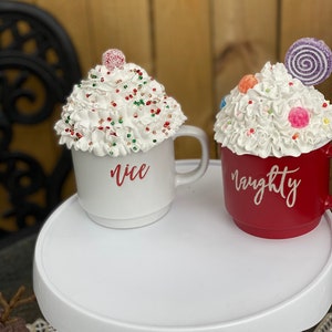 May include: Two ceramic mugs with white frosting and colorful sprinkles. One mug is white with the word "nice" in red lettering. The other mug is red with the word "naughty" in white lettering.