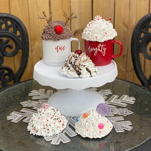May include: A white cake stand with three white and brown Christmas-themed hot chocolate mugs. The mugs are decorated with whipped cream, candy canes, and sprinkles. The mugs are labeled "nice" and "naughty".