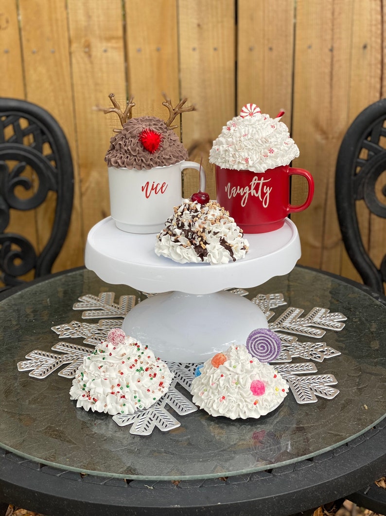 May include: A white cake stand with three white and brown Christmas-themed hot chocolate mugs. The mugs are decorated with whipped cream, candy canes, and sprinkles. The mugs are labeled "nice" and "naughty".