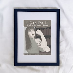 May include: A framed print with a black frame and a white background. The print features a woman with brown hair and a broken heart tattoo on her arm. The text on the print reads "I Can Do It With a Broken Heart" and "The Tortured Poets Department".