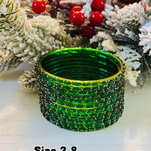 May include: A stack of green glass bangles with gold accents and small gold dots. The bangles are displayed in front of a festive background with red berries and white frosted branches. The text "Size 2.8" is visible.