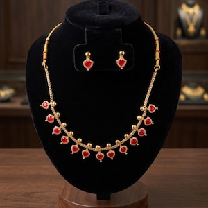 May include: A gold-toned necklace and matching earrings set displayed on a black velvet bust. The necklace features red heart-shaped pendants and small gold beads. The earrings have a similar design. The jewelry is presented on a wooden surface.