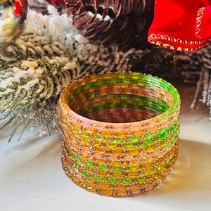 Pode incluir: Uma pilha de pulseiras coloridas em tons de verde, amarelo e rosa, adornadas com pequenos enfeites brilhantes. As pulseiras estão empilhadas sobre uma superfície branca, com decorações festivas de Natal ao fundo.