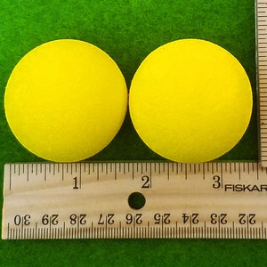 May include: Two bright yellow foam circles, each approximately 3 inches in diameter, are displayed on a green surface. A wooden ruler with inch markings is placed next to the circles for size comparison.