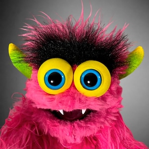 May include: A plush monster puppet featuring bright pink shaggy fur, a thick patch of black hair on top, and two large yellow eyes with blue pupils. The creature has pointed lime green ears and a wide mouth showing two small white fangs.