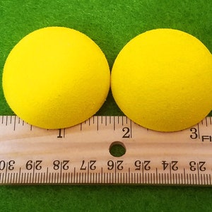May include: Two bright yellow foam balls, each about 1 inch in diameter, are placed next to each other on a wooden ruler. The ruler is marked with inches and fractions of an inch.