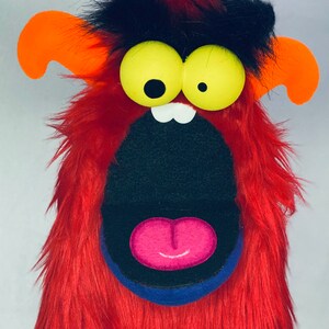 May include: A red furry puppet with large yellow eyes, a black mouth, and a pink tongue. The puppet has orange ears and a blue collar.