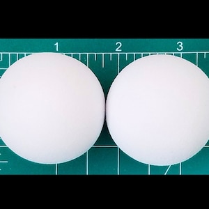 May include: Two white ping pong balls on a green cutting mat with inch markings.