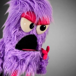 May include: A purple fuzzy monster puppet with long shaggy fur, large white eyes with black pupils, and bright pink eyelids. The puppet features a wide open black mouth, a pink tongue, and a small pink hand gesture visible on the side.