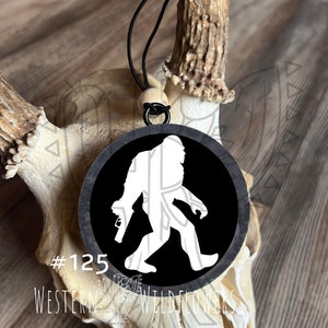 May include: A black and white round ornament with a white silhouette of Bigfoot holding a gun. The ornament is hanging from a black cord and a wooden bead. The ornament is hanging from a deer antler. The text #125 and WESTERN WILDFLOWERS is visible in the image.