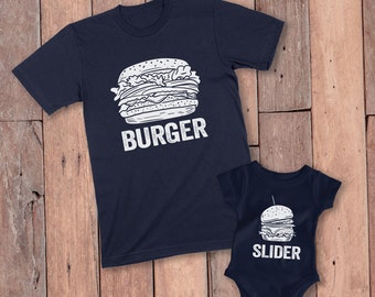 Può includere: Due capi blu navy con grafiche a tema burger. Una è una t-shirt con un grande hamburger e la scritta "BURGER". L'altro è un body per neonati con un hamburger più piccolo e la scritta "SLIDER". Sono esposti su una superficie di legno.