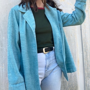 May include: A light blue blazer with a long sleeve and a single button closure. The blazer is worn over a dark green long-sleeve shirt and light blue jeans with a gold belt.