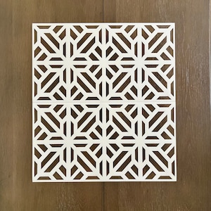 May include: White geometric stencil with a repeating pattern of intersecting lines creating a starburst design. The stencil is on a brown wood background.