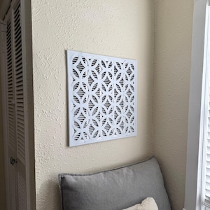 May include: A square white air vent cover featuring a repeating geometric pattern of interlocking circles. The cover is mounted on a textured beige wall, positioned above a gray cushion, and displays a series of horizontal slats behind the design.