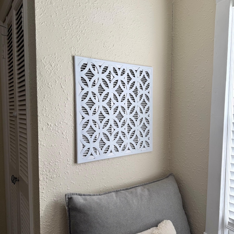 May include: A square white air vent cover featuring a repeating geometric pattern of interlocking circles. The cover is mounted on a textured beige wall, positioned above a gray cushion, and displays a series of horizontal slats behind the design.