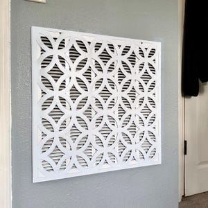 May include: A square white air vent cover featuring a repeating geometric pattern of interlocking circles. The decorative grille is mounted flush against a gray wall, with horizontal slats visible behind the intricate white cutout design.
