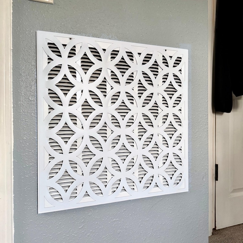 May include: A square white air vent cover featuring a repeating geometric pattern of interlocking circles. The decorative grille is mounted flush against a gray wall, with horizontal slats visible behind the intricate white cutout design.