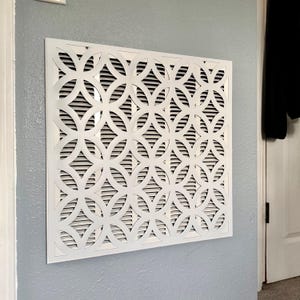 May include: White decorative air vent cover with a geometric pattern of interlocking circles and curved lines. The cover is mounted on a light blue wall. The vent has horizontal slats behind the decorative overlay.