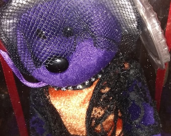 May include: A purple plush bear wearing a black lace veil and a black lace shawl over an orange dress. The bear has a black button nose and a beaded necklace. The bear is in a clear plastic display case.