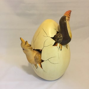 May include: A ceramic sculpture of a toucan and a chick emerging from a cracked, pale yellow egg. The toucan is brown with a yellow beak and the chick is yellow.