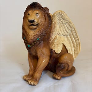May include: A winged lion figurine with a brown and tan color scheme. The lion is sitting with its wings spread out behind it. It has a gold chain with turquoise gems around its neck.