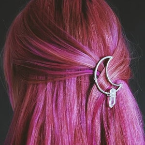 Pode incluir: Um grampo de cabelo de prata em forma de lua crescente com um cristal transparente pendurado na parte inferior. O grampo está no cabelo rosa.