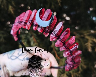 May include: A headband featuring a series of raw, deep red crystal points secured with thin black wire. A silver-colored crescent moon charm is attached to the center. The logo for Luna Corvus, depicting a raven above crystals, is visible.