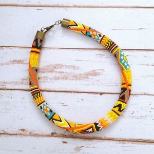 May include: A beaded necklace with a vibrant, geometric pattern. The necklace features a yellow base with orange, black, white, and turquoise accents. The necklace is approximately 18 inches in length and has a brass clasp.