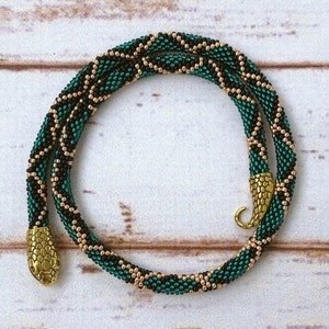 May include: A beaded necklace in teal, black, and gold, featuring a snake-like design. The necklace has a textured pattern and gold-colored snake head clasps. The jewelry is displayed on a light-colored wooden surface.