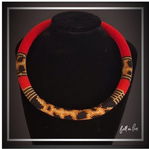 May include: A red beaded necklace with a black and gold leopard print design. The necklace is displayed on a black mannequin. The necklace is a choker style.
