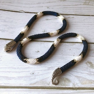 May include: A beaded necklace with a snake design. The necklace is made of blue, white, and gold beads. The necklace has a silver clasp in the shape of a snake head.