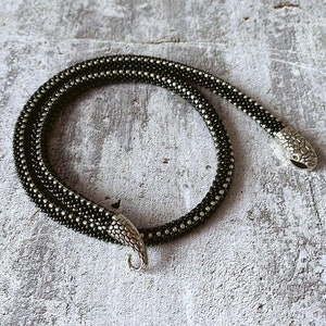 May include: A black and silver beaded necklace with a snake-like design. The necklace is made of small, round beads and features silver-toned snake head clasps. The necklace is displayed on a textured, gray surface.