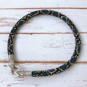 May include: A circular beaded bracelet featuring a repeating snake-like pattern in black, iridescent blue, and silver beads. The bracelet is finished with two silver-toned metal snake head end caps that connect with a circular silver-toned clasp.