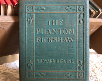 Pode incluir: Um livro de capa dura na cor verde-azulado intitulado The Phantom Rickshaw de Rudyard Kipling. A capa apresenta uma borda retangular em relevo com motivos florais nos cantos. O título e o autor estão impressos em uma fonte escura.