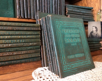 May include: A collection of dark green hardcover books arranged on a wooden shelf. The front-most book features the title "Friendship and Other Essays" by Henry Thoreau in embossed lettering, framed by a decorative border design.