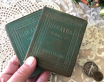 Pode incluir: Dois livros pequenos e desgastados de cor verde-azulada com capas em relevo. O texto na capa diz HIAWATHA VOL. I e LONGFELLOW. Os livros apresentam padrões decorativos nas bordas e uma textura gasta. As capas são de um tom verde suave.